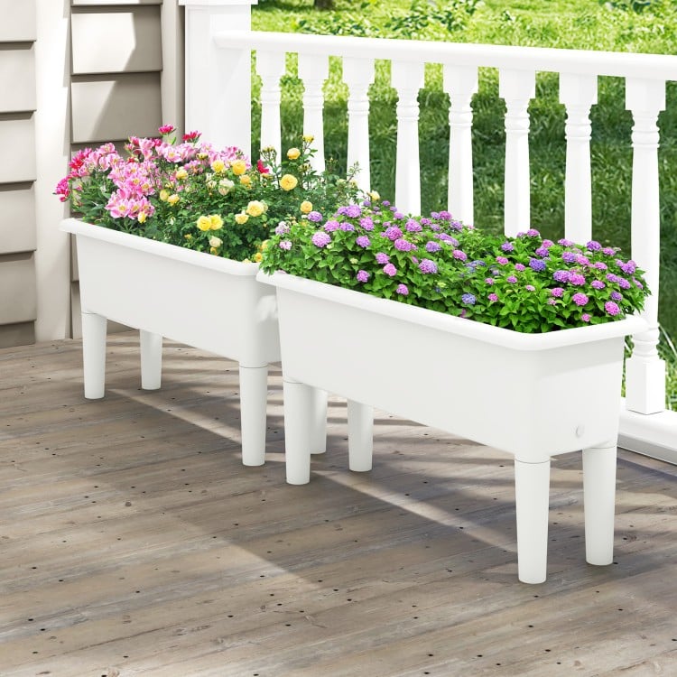 Two white self-watering planter boxes with flowers, featuring detachable legs and drainage holes for outdoor gardening.
