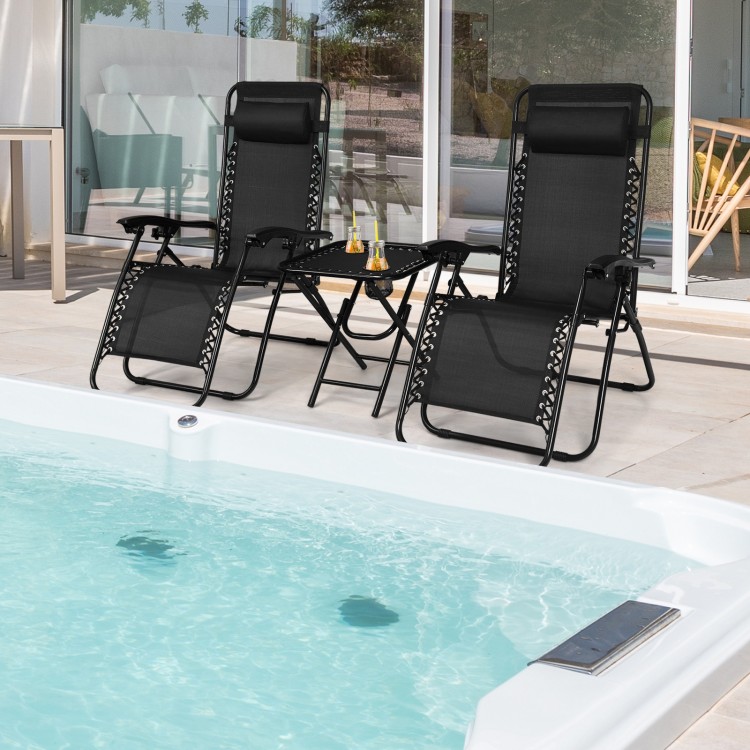 Two black folding zero gravity reclining lounge chairs with a table, perfect for outdoor relaxation by the pool.