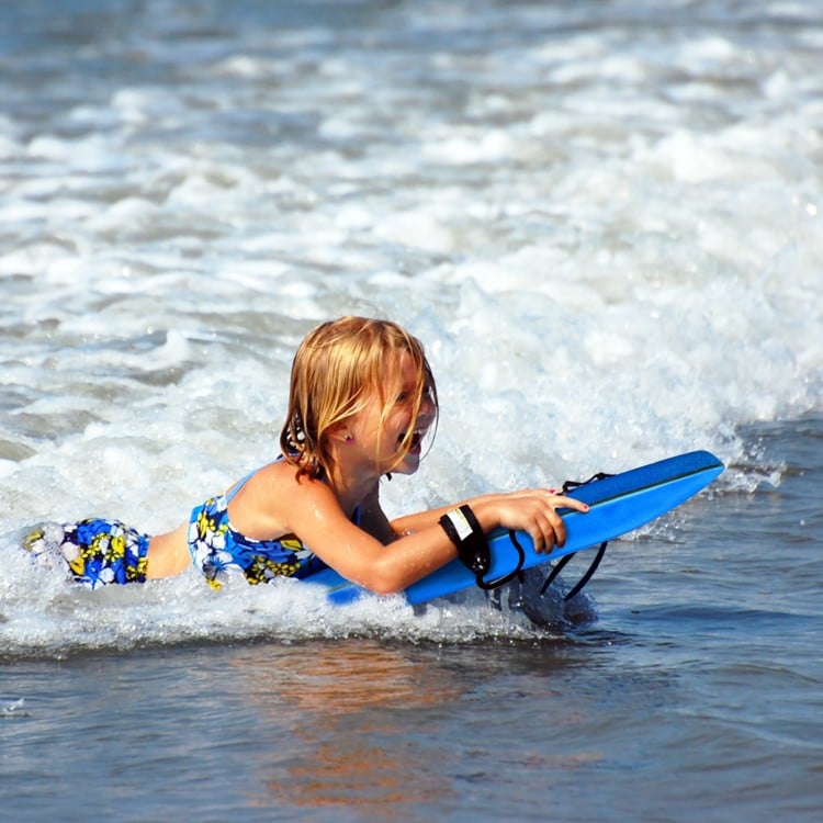 Child riding a blue bodyboard in the ocean, featuring an EPS core and crescent tail for enhanced surfing experience.