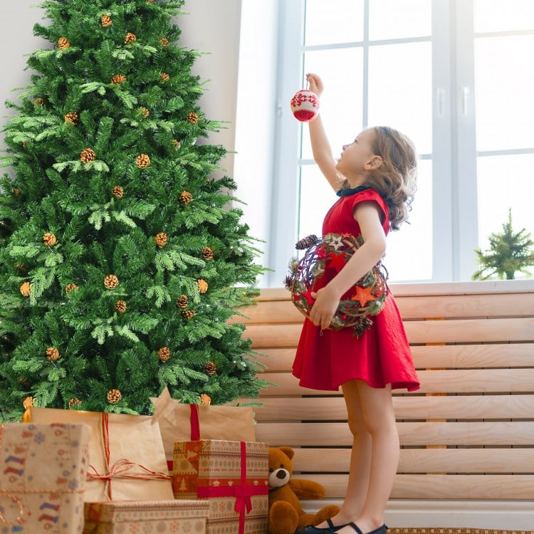 Child in a red dress decorates a 7' artificial Christmas tree with LED lights and pine cones for festive indoor.