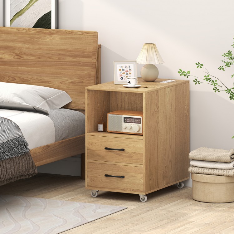 Natural wood end table with charging station, two drawers, and wheels for easy mobility beside a bed.
