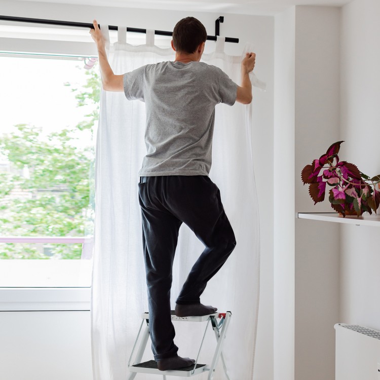 A person using a folding 2 step ladder with anti-slip pedals to hang curtains in a bright room.