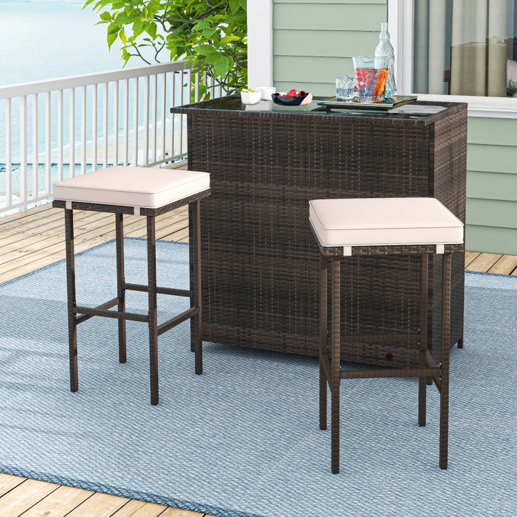 Patio bar set featuring a brown rattan table with tempered glass top and two cushioned stools for outdoor relaxation.