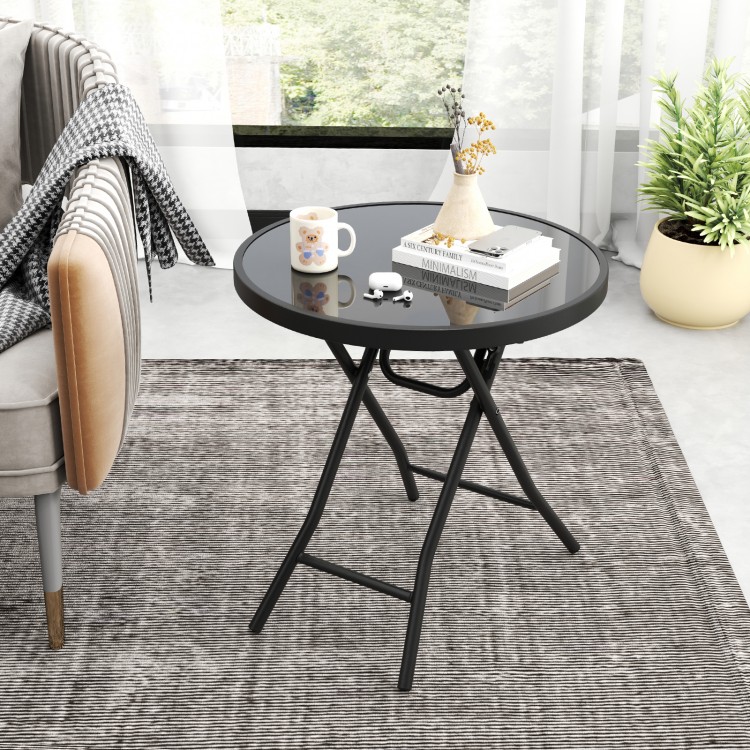 Round black folding side table with tempered glass top, perfect for indoor and outdoor use, shown in a living room.