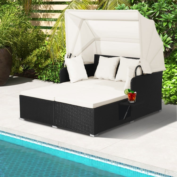 Patio rattan daybed with retractable canopy, off-white cushions, and side tables, perfect for poolside relaxation.