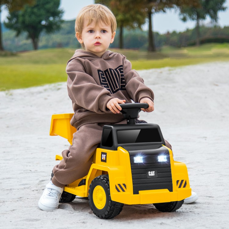 Child riding a yellow 6V electric Caterpillar construction vehicle with detachable dump bed and LED lights on a sandy.
