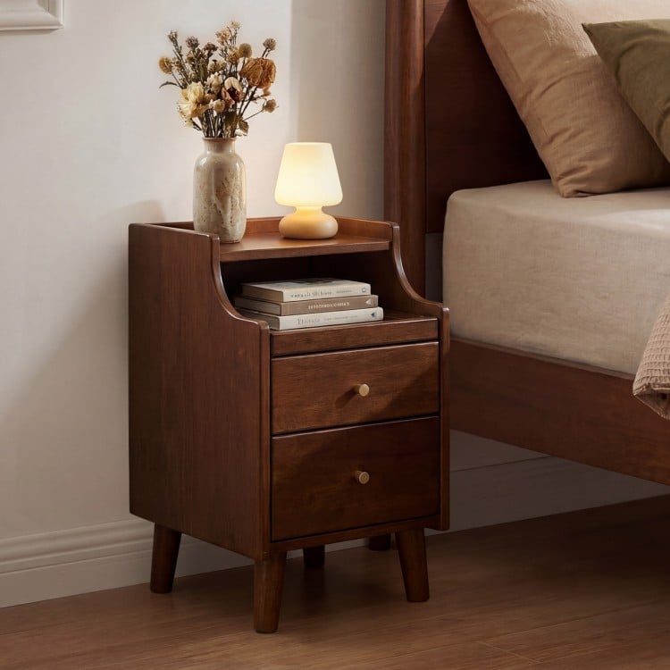 Brown wood nightstand with two drawers, solid wood legs, and a pull-out tray for cable management beside a bed.