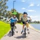 Two kids riding 12-inch white bikes with training wheels, enjoying a smooth ride along a scenic path by the water.