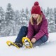 A child wearing a purple jacket and red hat adjusts yellow 17” kids snowshoes in snowy terrain for winter fun.