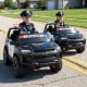 Two kids in a black 24V 2-seater ride-on police car with LED lights, designed for ages 3 to 8, on a road.