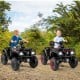 Two kids riding a 24V black ATV 2-seater with LED lights and music on a dirt path, perfect for outdoor fun.