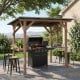 Grill gazebo with cedar wood frame, sloped metal roof, and two side serving shelves for outdoor BBQ gatherings.