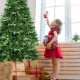 Child in a red dress decorates a 7' artificial Christmas tree with LED lights and pine cones for festive indoor.