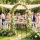 Heavy-duty black metal garden arch with gate, adorned with flowers, ideal for weddings and outdoor events.