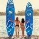 Two people holding blue inflatable stand-up paddle boards by the water, perfect for surfing and yoga.