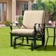 Patio glider rocking chair with thick tan cushion and curved armrests, perfect for outdoor relaxation.