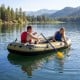 Three-person inflatable fishing raft in green, featuring paddlers on a lake with mountains in the background.