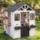 Kids playing in a white wooden playhouse with a sink, flower pots, and a half door for outdoor fun and imaginative play.