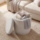 Round beige storage ottoman with removable lid, acacia wood frame, and cozy blankets inside for versatile use.