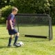 Youth player kicking a soccer ball towards a 6 ft x 4 ft metal frame goal post in a backyard setting.